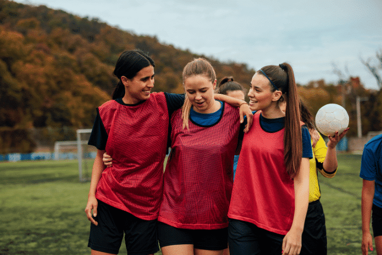 How Do Football Players Communicate On and Off the Field - Three different female football players bonding on a soccer pitch